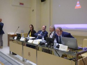 Cinque relatori al tavolo di una sala conferenze, con i cartellini dei nomi e i microfoni davanti.
