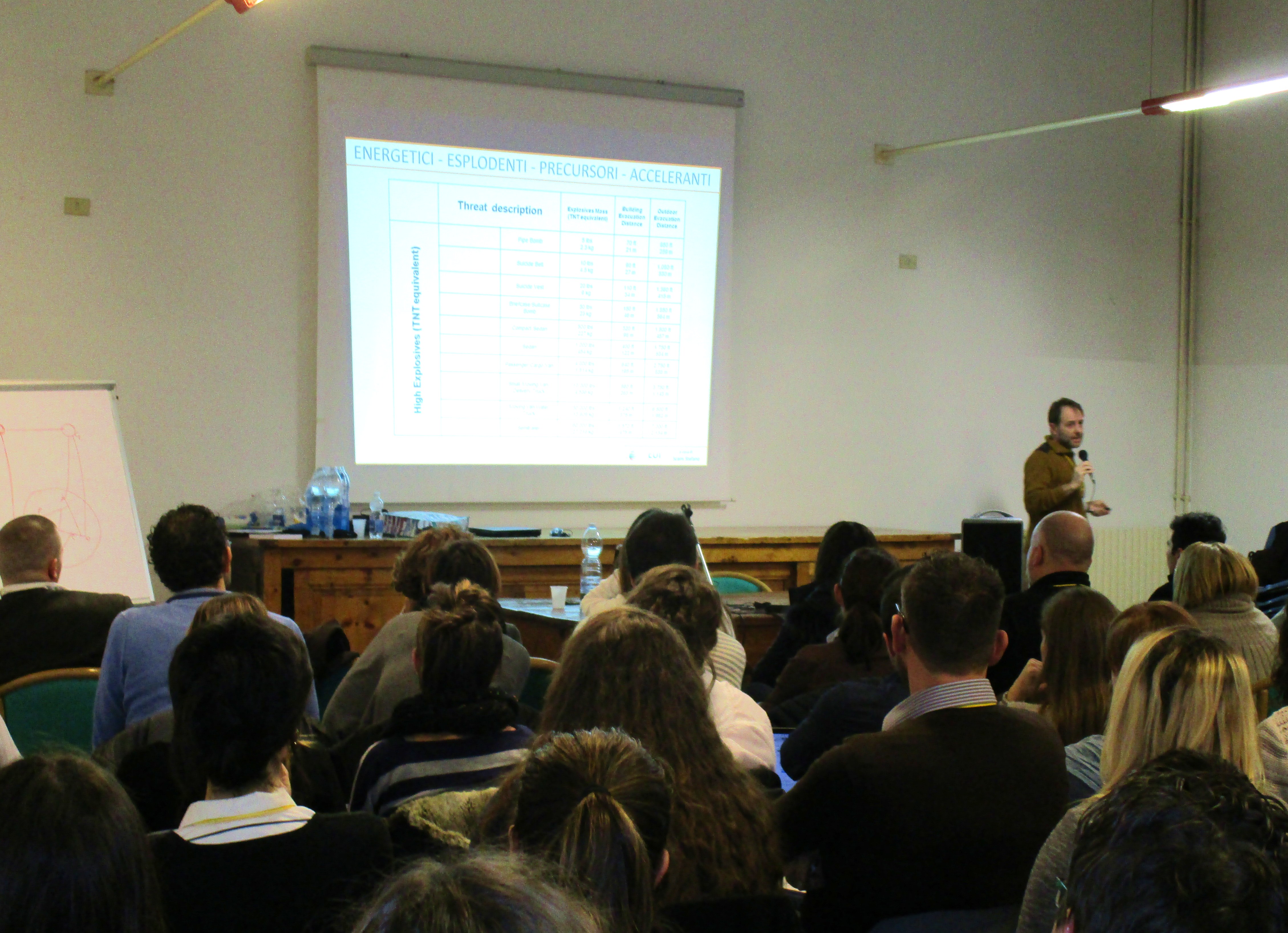 Un relatore accanto alla proiezione di una tabella di dati, davanti all'aula piena.