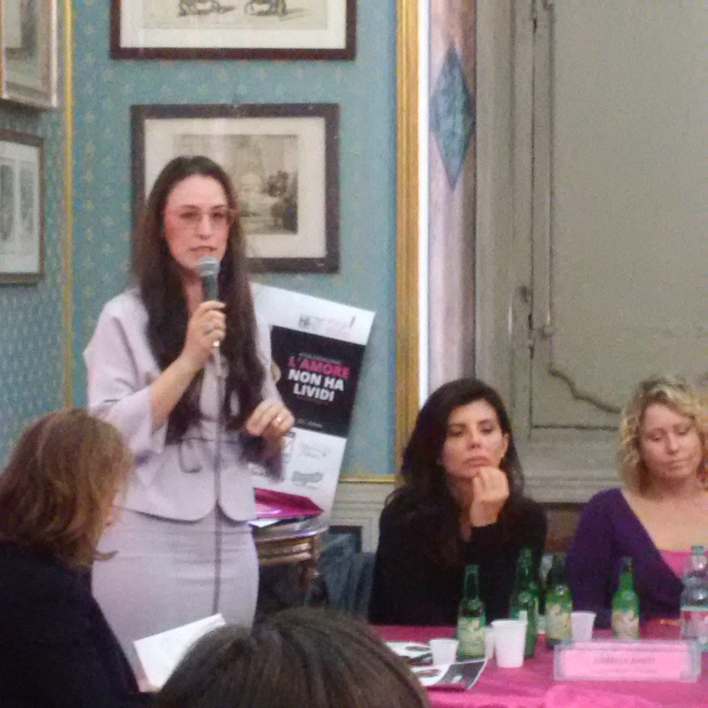 Una relatrice al microfono mostra il manifesto L'amore non ha lividi alle altre partecipanti.