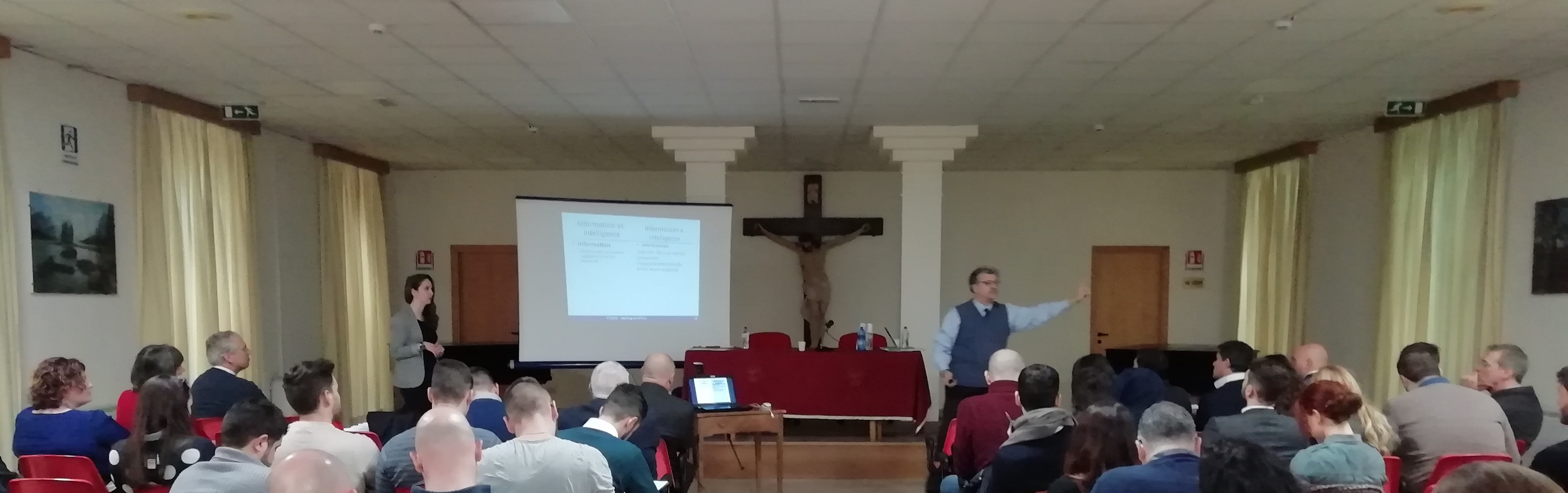Panoramica della sala durante la lezione: il pubblico seduto e due relatori accanto allo schermo.