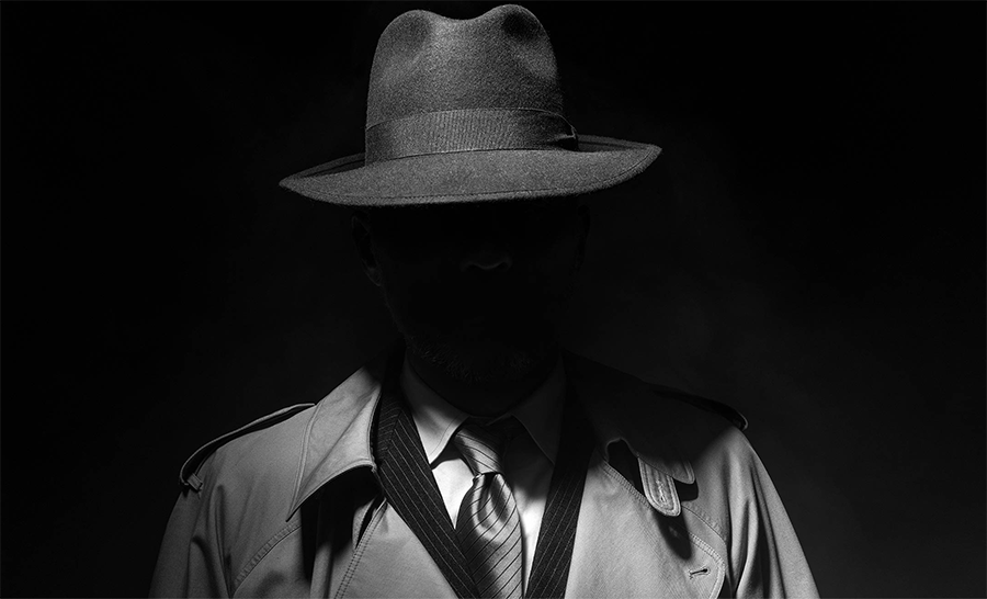 Fotografia in bianco e nero: un uomo in impermeabile e cappello a falda larga, il viso nell'ombra.