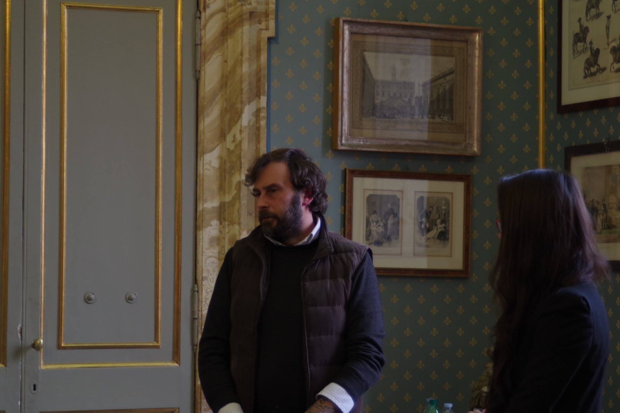 Due persone in piedi parlano fra le stampe appese alla parete di una sala storica.