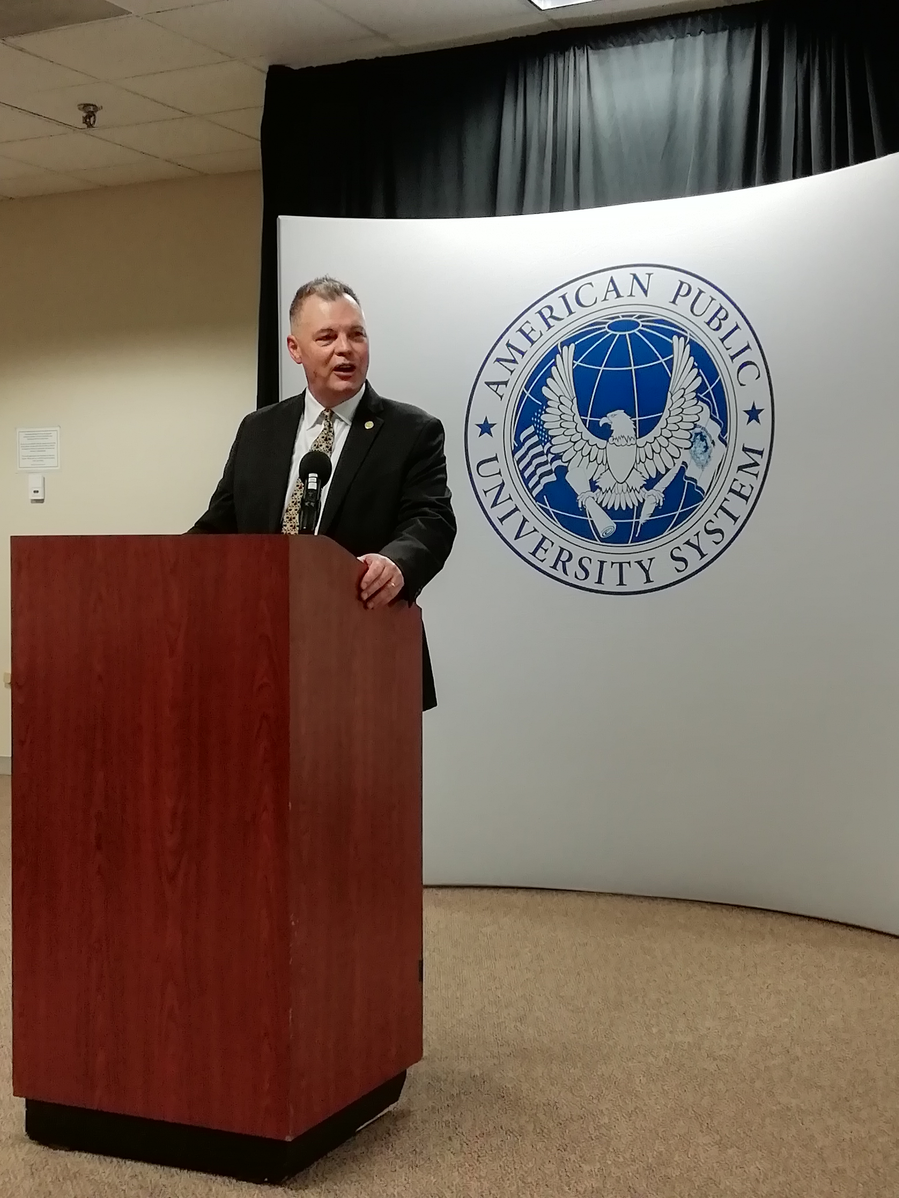 Un relatore parla dal podio accanto al pannello con il sigillo dell'American Public University System.