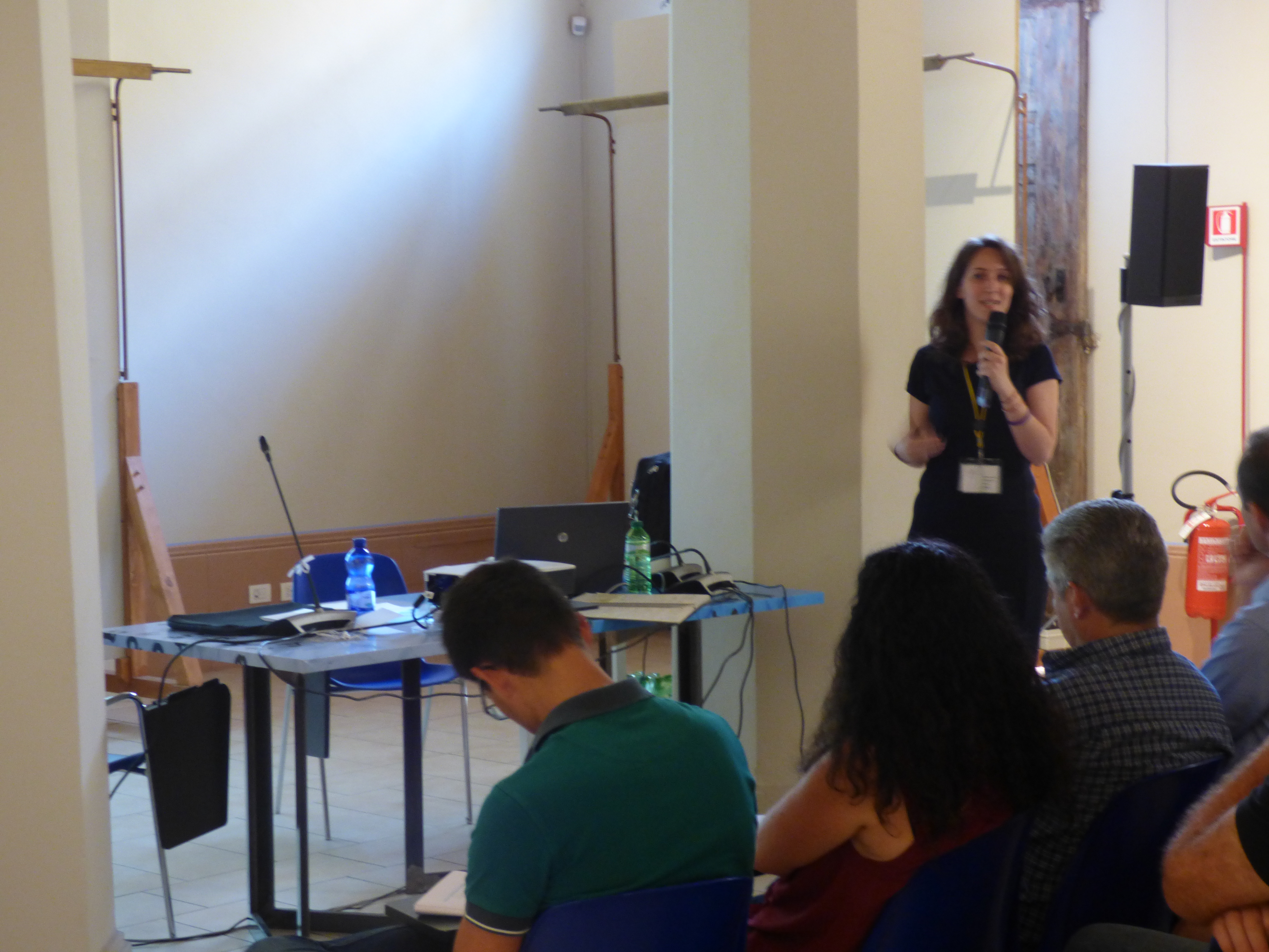 Una relatrice al microfono accanto al tavolo dei computer, davanti al pubblico.