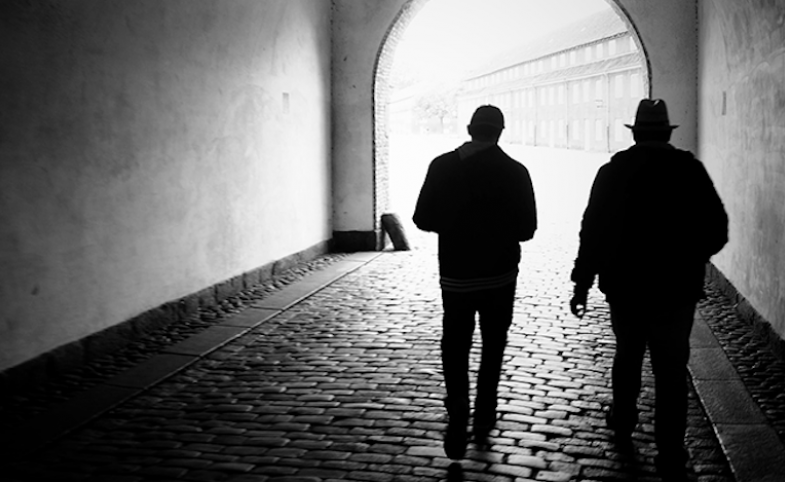 Fotografia in bianco e nero: due uomini di spalle, in controluce, camminano lungo un passaggio coperto di ciottoli.