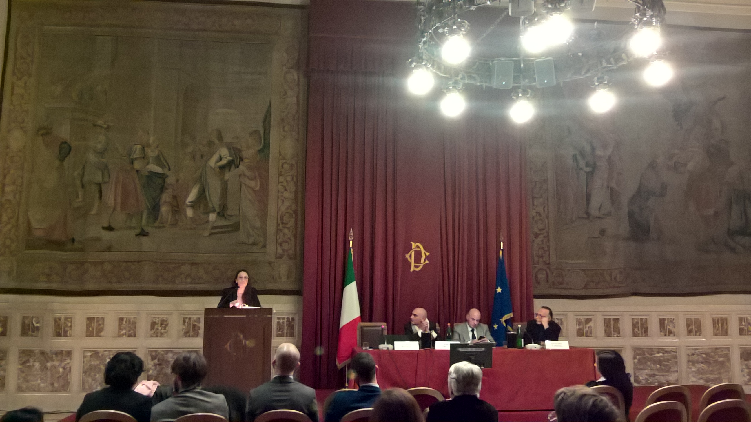 Una relatrice parla dal podio di una sala parlamentare affrescata, accanto alla bandiera italiana e a quella europea; al tavolo tre persone.