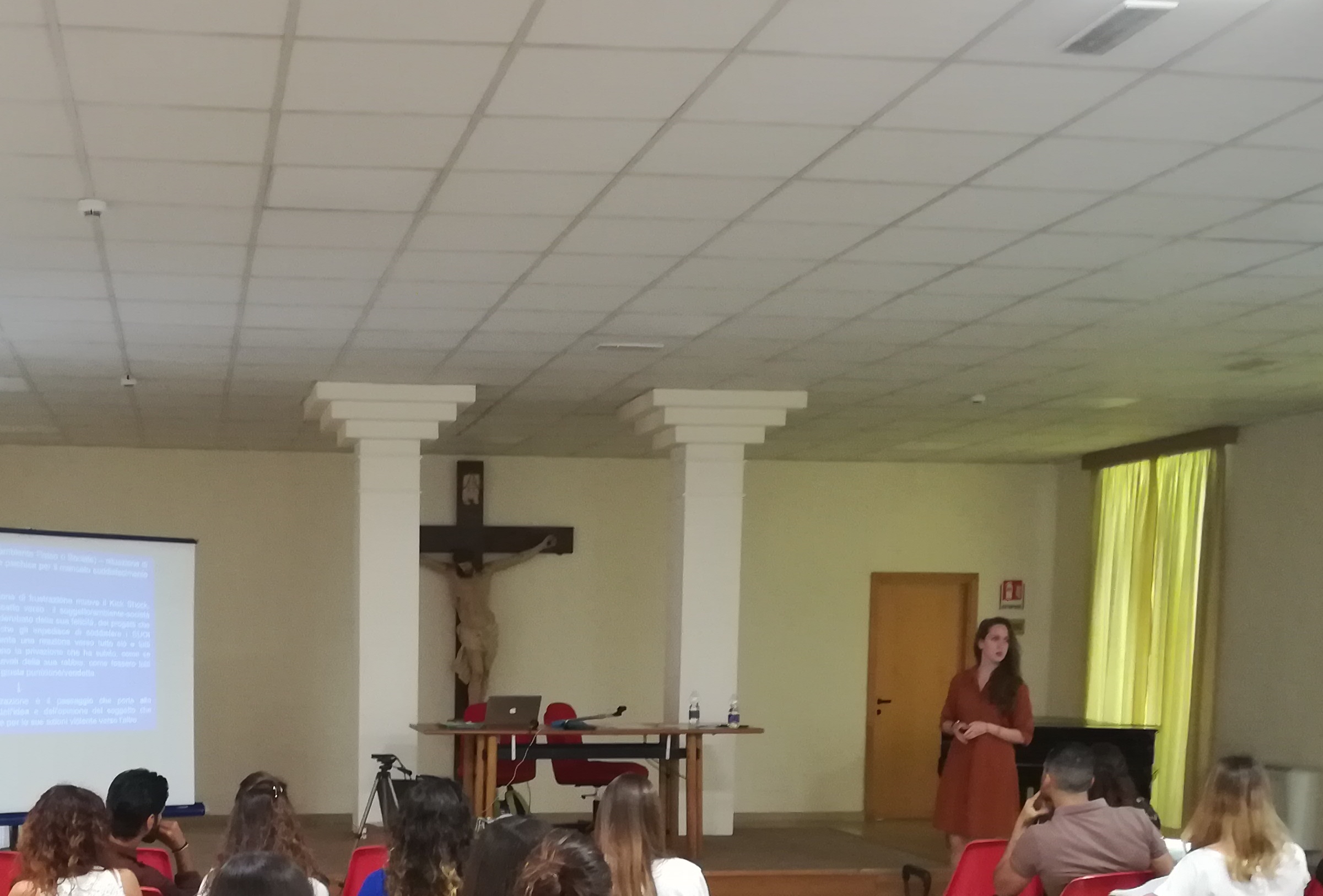 Una relatrice accanto alla proiezione, in una sala con il crocifisso e le colonne.