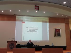 Un relatore al tavolo di un'aula circolare, sotto la proiezione di una definizione di intelligence.