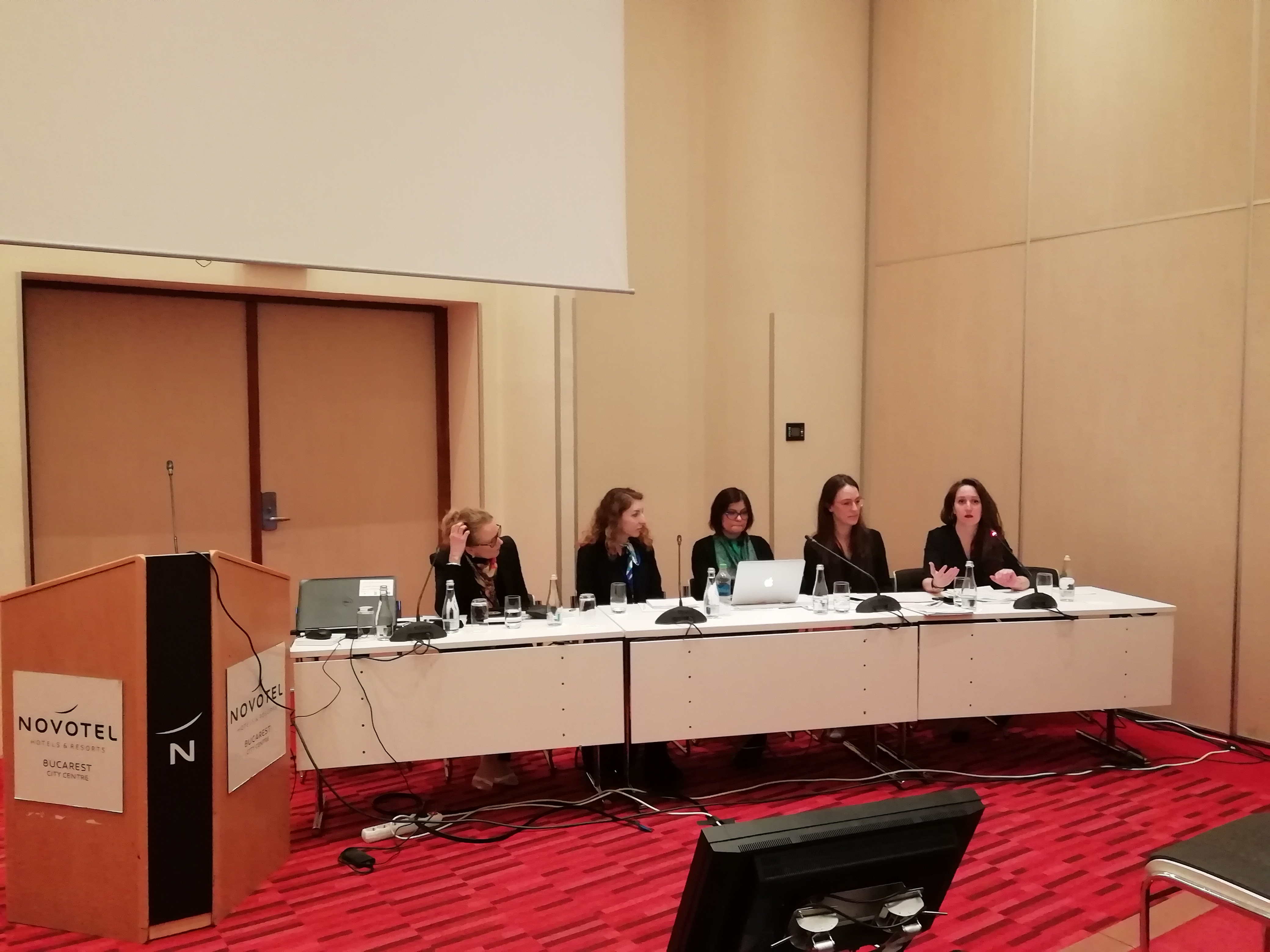 Cinque relatrici al tavolo dei lavori, davanti al podio con la scritta Novotel.