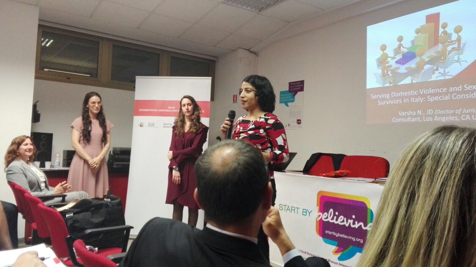 Una relatrice parla al microfono fra il pubblico, con altre tre relatrici in piedi accanto al banner.