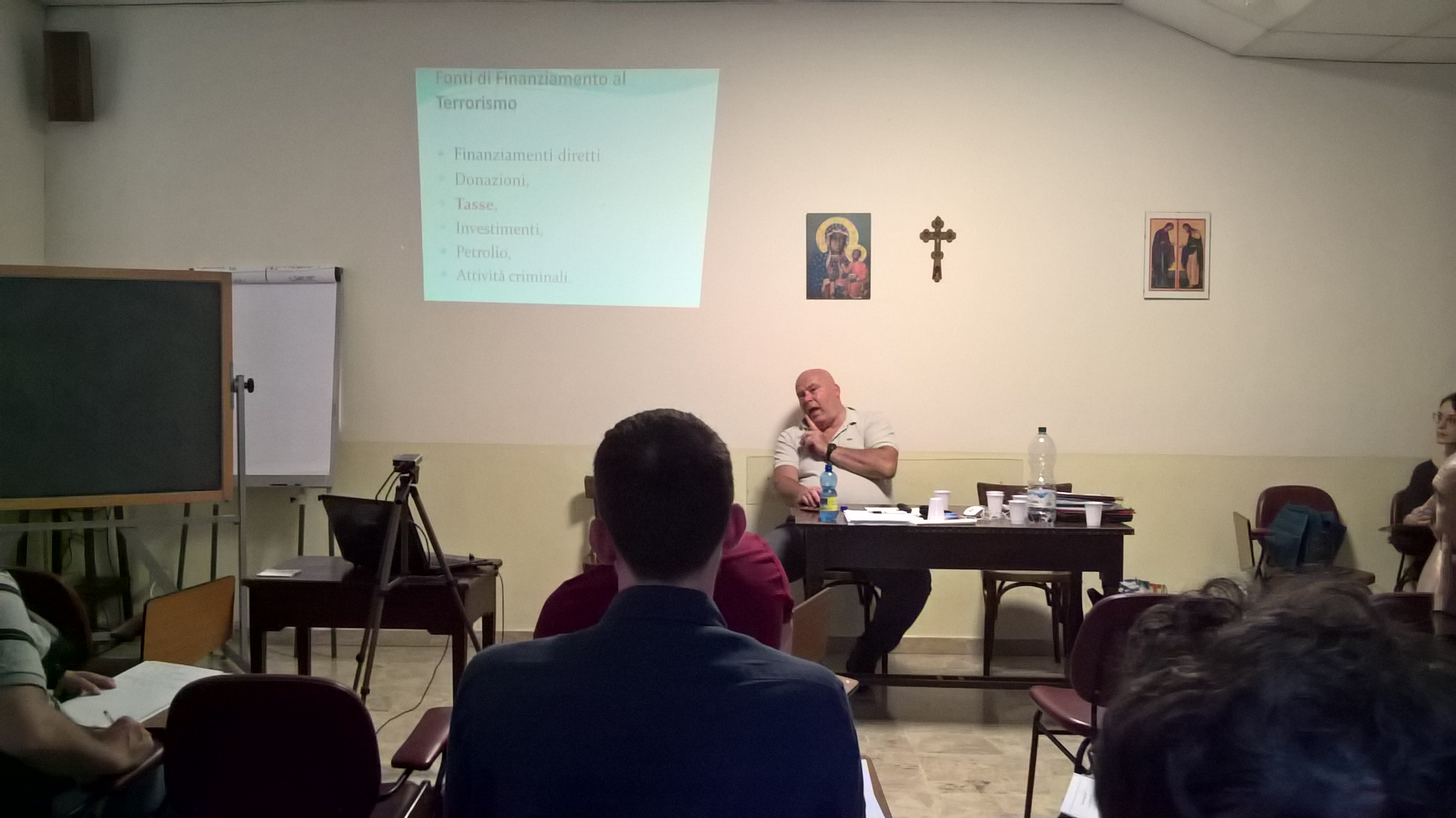 Un relatore seduto al tavolo parla accanto alla proiezione, sotto un crocifisso appeso al muro.